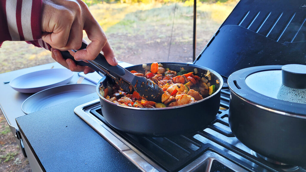 A person is stirring a recipe of harissa spiced vegetables and couscous with tongs over a camp stove.