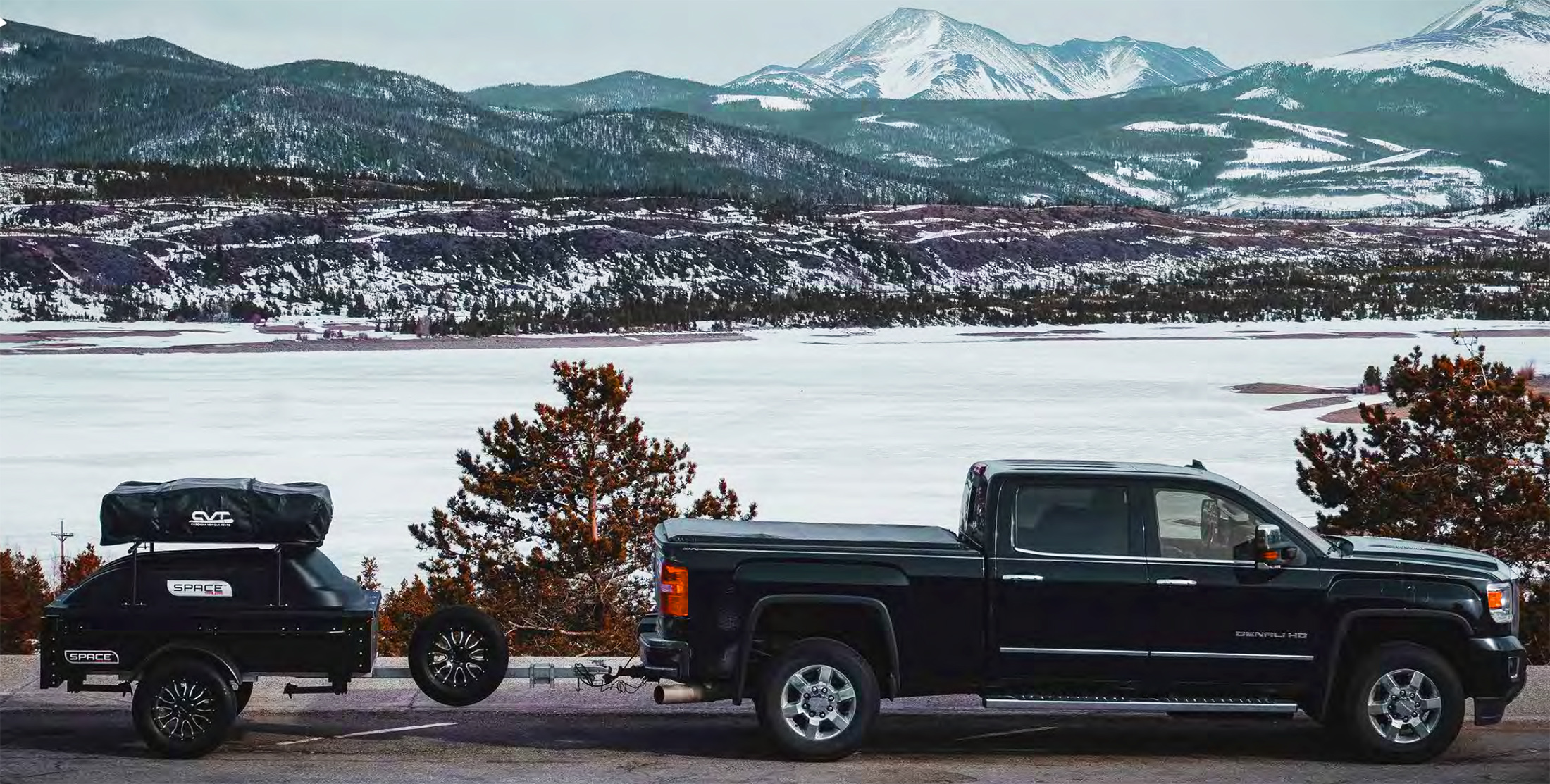 A closed SPACE trailer is shown hitched behind a green truck in winter, with snow capped mountains in the background