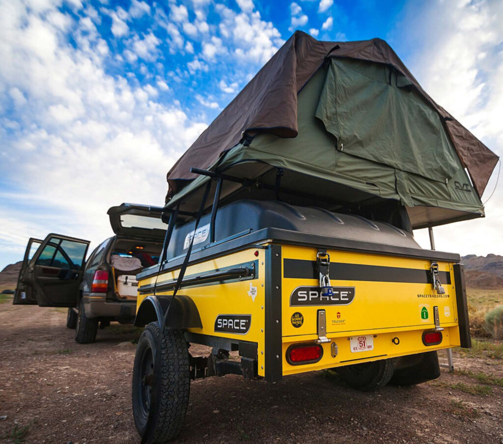 A yellow SPACE trailer is shown behind a vehicle on a sandy area, with the tent set up above it