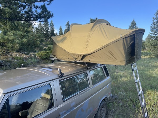 Thule Approach M rooftop tent in beige, set up on a van in a field that borders a forest.